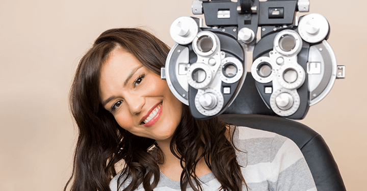 A smiling woman at an eye exam with an optometrist and seated with a phoropter optometric instrument